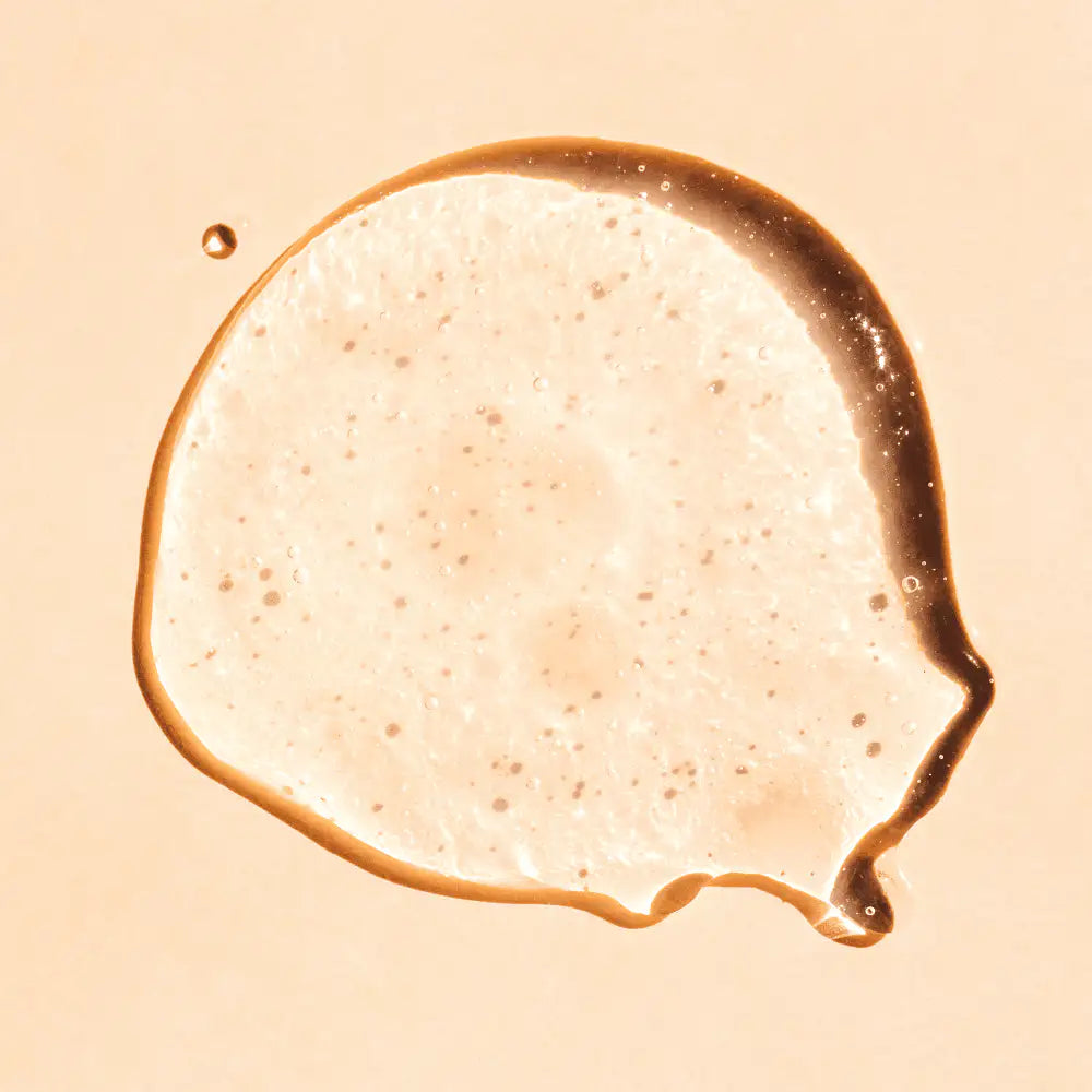 A glossy droplet of translucent beige cosmetic serum or oil with fine speckles, shown on a warm beige background.