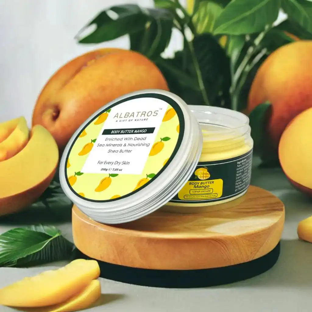 Albatros 100% Butter Mango Enriched Body Butter jar with its lid leaning on a wooden stand, surrounded by fresh mango slices, whole mangoes, and green leaves.